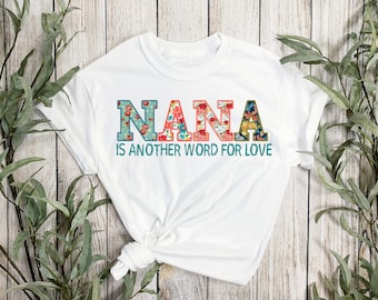May include: White t-shirt featuring the word "NANA" in floral print lettering. The phrase "IS ANOTHER WORD FOR LOVE" is printed below in teal. The shirt is displayed against a light wood backdrop, with green leafy branches.