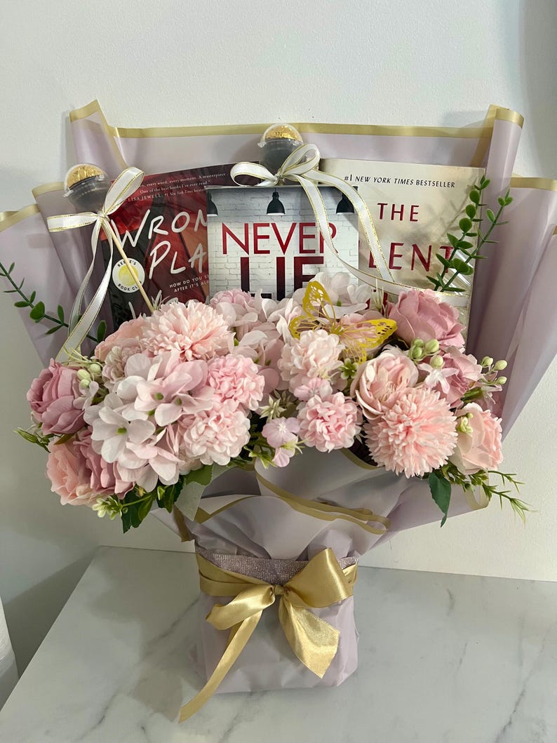 A bouquet of pink flowers wrapped with lilac and gold tissue paper and a gold bow, holding three different books in the bouquet