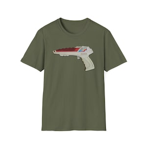 Puede incluir: Camiseta verde oliva con un gráfico de una pistola de rayos futurista en blanco, rojo y azul. El diseño de la pistola de rayos está en el lado izquierdo de la camiseta. La camiseta tiene cuello redondo y mangas cortas.