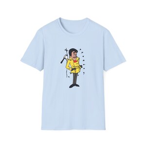 Può includere: Maglietta blu chiaro con un'illustrazione di un uomo cartone animato con un cappotto giallo e baffi che tiene un bastone. L'uomo è in piedi di fronte a un muro con punti neri.