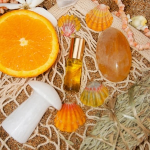 Op de afbeelding: Een arrangement van natuurlijke elementen op zand. Bevat een sinaasappelschijf, schelpen, een heldere kwartskristal, een kleine oliefles, een wit paddenstoelvormig object en een groot oranje kristal. Een net en een bundel gedroogde kruiden zijn ook aanwezig.
