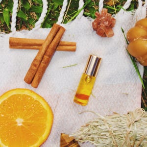 Op de afbeelding: Een arrangement van natuurlijke elementen op een witte doek, waaronder kaneelstokjes, een sinaasappelschijf, een kleine rollerfles met essentiële olie, een saliebundel en een tijgeroogkristal. Op een witte doek met groene grasachtergrond.