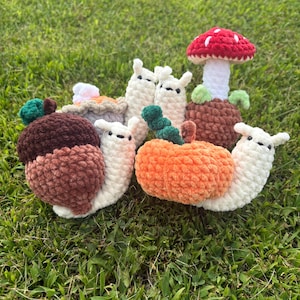 May include: A collection of crocheted plush figures including snails with shells shaped like an acorn, a pumpkin, and a pie, alongside a small white snail and a red-capped mushroom with a white stem and green leaf accents.