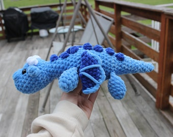 May include: A handmade, plush crochet dinosaur in shades of blue. The toy features a textured body, dark blue spines, and a white accent on its head. The dinosaur is held in front of a wooden deck.