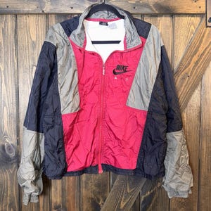May include: A color-blocked Nike Air windbreaker jacket featuring a vibrant red center panel, dark navy blue side panels, and muted olive green accents on the sleeves and chest. The jacket has a full-length front zipper and a black Nike Air logo.