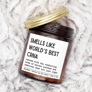 May include: A brown glass jar candle with a gold lid, resting on a fluffy white surface. The label reads "SMELLS LIKE WORLD'S BEST CRNA" with additional text about the scent. The candle is from WockoWicks and made in the USA.