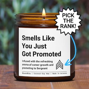 May include: A brown glass candle with a white label that reads "Smells Like You Just Got Promoted". The label also includes the text "Infused with the refreshing aroma of career growth and promoting to Sergeant" and a black and white image of three chevrons. The candle is on a wooden surface.