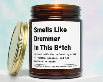 May include: A brown glass jar candle with a gold lid, labeled "Smells Like Drummer In This B*tch." The label also states the candle is infused with the aroma of rhythm, passion, and music. The lid is off to the side.