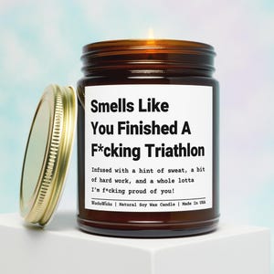May include: A brown glass jar candle with a gold lid, labeled "Smells Like You Finished A F*cking Triathlon." The label includes text about sweat, hard work, and pride. The candle is a natural soy wax candle.