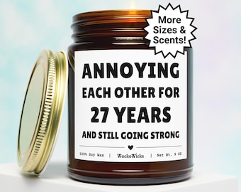 May include: A lit brown glass jar candle with a gold lid, labeled "ANNOYING EACH OTHER FOR 27 YEARS AND STILL GOING STRONG." The candle is 9 oz and made of 100% soy wax. A white starburst graphic reads "More Sizes & Scents!"
