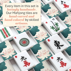 May include: A collection of white Mahjong tiles featuring hand-carved and hand-colored designs of pink lotus flowers, green leaves, bamboo, and black or red Chinese characters. Text on the image reads: Every item in this set is lovingly handmade. Our Mahjong tiles are hand-carved and hand-colored by skilled artisans.