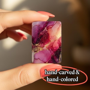 May include: A rectangular piece with an abstract alcohol ink design in shades of magenta, deep purple, and soft pink, accented with gold flecks. The surface features the text hand-carved & hand-colored in a black serif font at the bottom right.