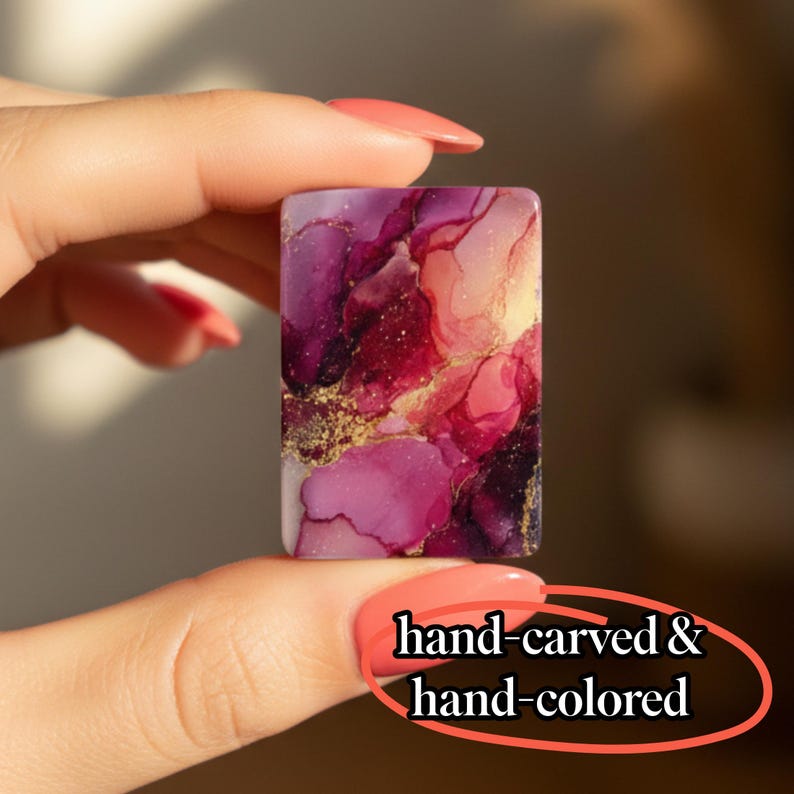May include: A rectangular piece with an abstract alcohol ink design in shades of magenta, deep purple, and soft pink, accented with gold flecks. The surface features the text hand-carved & hand-colored in a black serif font at the bottom right.