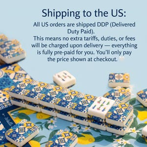 May include: A set of mahjong tiles featuring a blue, yellow, and white geometric tile pattern on the back. The front of one tile displays a green bamboo design with the number seven. The tiles are arranged on a surface with a lemon and floral print.