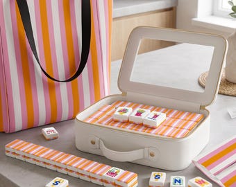 May include: A mahjong set featuring tiles with orange and pink striped backs, stored in a white rectangular carrying case with a clear window. A matching striped tote bag, a long row of tiles, and several individual tiles with colorful icons are included.