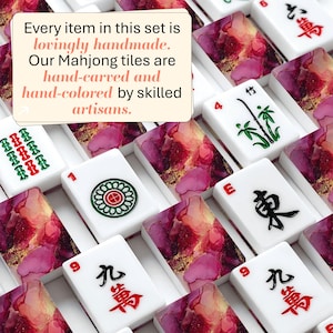 May include: A collection of Mahjong tiles featuring white surfaces with black, red, and green ink symbols and backs with a pink, purple, and gold marbled pattern. Text on the image reads: Every item in this set is lovingly handmade. Our Mahjong tiles are hand-carved and hand-colored by skilled artisans.