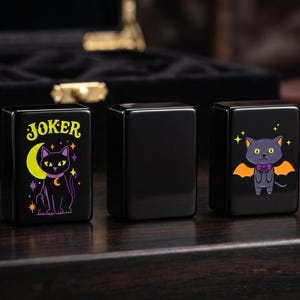 May include: Three rectangular black tiles with a glossy finish. The left tile features a yellow crescent moon and a black cat with the word JOKER printed above. The middle tile is blank. The right tile displays a gray cat with orange bat wings.
