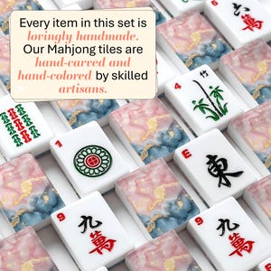 May include: A collection of rectangular Mahjong tiles featuring white surfaces with black, red, and green character engravings, interspersed with tiles displaying a pink, blue, and gold marbled pattern. Text reads: Every item in this set is lovingly handmade. Our Mahjong tiles are hand-carved and hand-colored by skilled artisans.