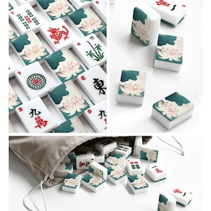 May include: A collection of white mahjong tiles featuring various red, green, and black ink illustrations including lotus flowers, bamboo, dragons, and Chinese characters. Some tiles are arranged in a grid, while others are scattered near a bag.