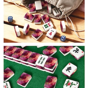May include: A set of mahjong tiles featuring a white base with a purple, pink, and gold marbled pattern on the back. The tiles display various traditional Chinese characters, symbols, and numbers in black, red, and green ink, accompanied by two blue dice.