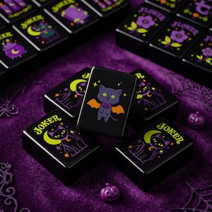May include: A set of black rectangular mahjong tiles featuring colorful illustrations of cats, bats, flowers, and the word JOKER in yellow. The tiles display purple and orange accents, with some tiles showing a cat with bat wings and others a cat with a moon.
