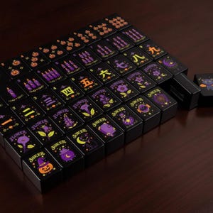 May include: A set of black mahjong tiles featuring Halloween-themed illustrations in purple, orange, and yellow. The tiles display various designs including jack-o'-lanterns, candles, flowers, cats, and potions, alongside traditional Chinese numerals.