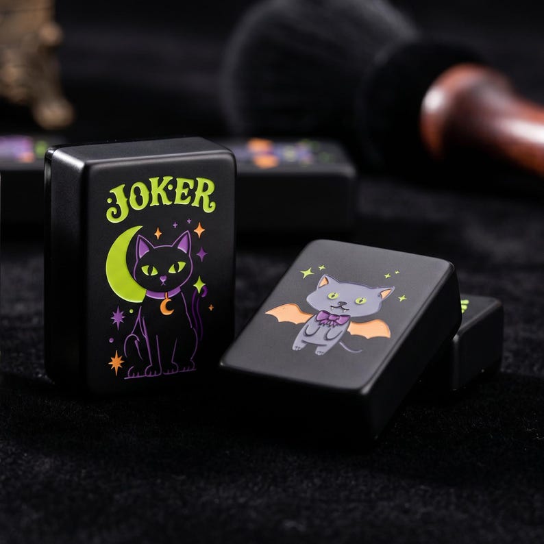 May include: Two rectangular black tiles featuring graphic illustrations. One tile displays a black cat sitting before a lime green crescent moon with the word JOKER in green text. The other tile shows a grey cat with orange bat wings and a purple bow.