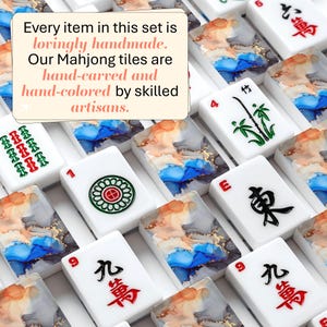 May include: A collection of white Mahjong tiles featuring traditional red, green, and black characters alongside tiles with abstract blue, orange, and gold ink patterns. A text box reads: Every item in this set is lovingly handmade. Our Mahjong tiles are hand-carved and hand-colored by skilled artisans.