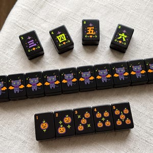 May include: A set of black rectangular mahjong tiles featuring Halloween-themed illustrations. Some tiles display purple cats with orange bat wings, others show orange jack-o'-lanterns, and four tiles feature green and yellow Chinese characters.