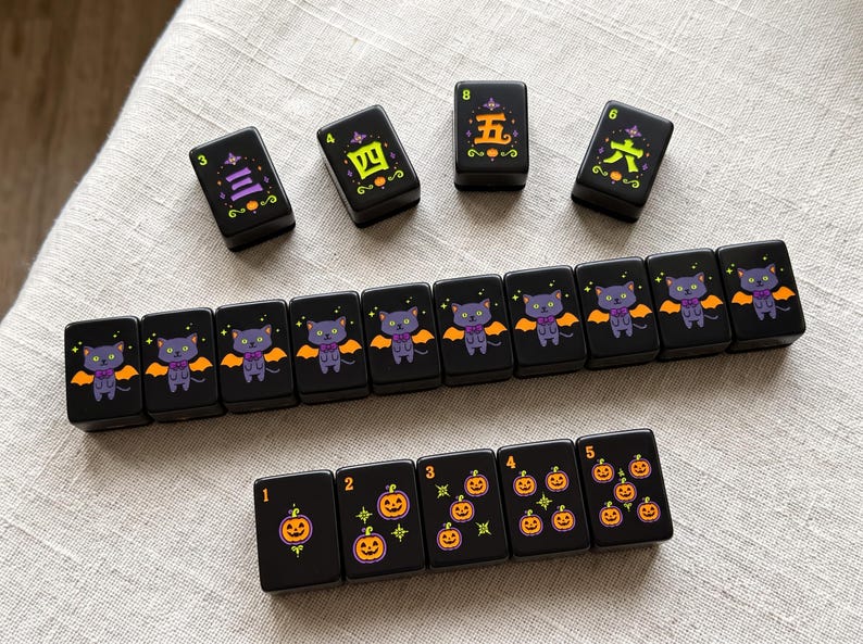 May include: A set of black rectangular mahjong tiles featuring Halloween-themed illustrations. Some tiles display purple cats with orange bat wings, others show orange jack-o'-lanterns, and four tiles feature green and yellow Chinese characters.