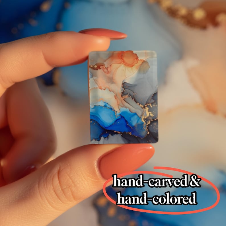 May include: A rectangular piece featuring an abstract alcohol ink design with swirls of blue, grey, peach, and gold metallic accents. The surface displays a smooth finish with the text hand-carved & hand-colored visible in the lower right corner.