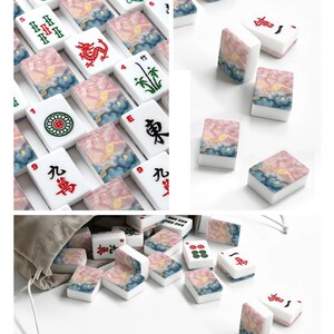 May include: A collection of rectangular Mahjong tiles featuring white faces with black, red, and green traditional symbols and characters. The backs of the tiles display a marbled pattern in shades of pink, blue, and gold. A beige drawstring bag is included.