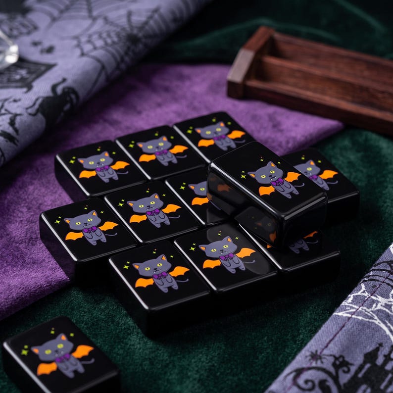 May include: A collection of rectangular black tiles featuring a cartoon illustration of a grey cat with orange bat wings and a purple bow tie. Each tile displays the same graphic of the cat with small yellow stars surrounding it on a glossy surface.