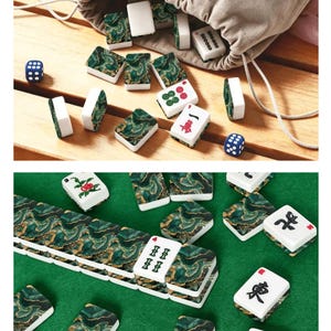 May include: A mahjong set featuring rectangular tiles with a green and gold marbled pattern on the back and white faces with various black, red, and green symbols. The set includes two blue six-sided dice and a beige drawstring storage bag.