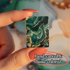 May include: A rectangular tile featuring a dark green and black marbled pattern with gold veining accents. The surface displays the text hand-carved & hand-colored. The object is held between two fingers, showcasing its small, flat, and smooth design.