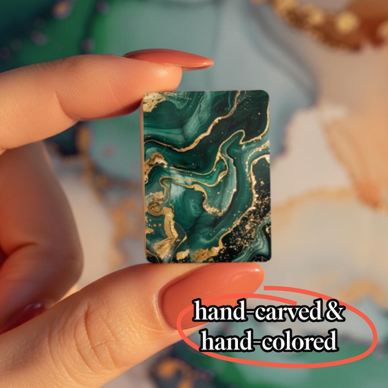 May include: A rectangular tile featuring a dark green and black marbled pattern with gold veining accents. The surface displays the text hand-carved & hand-colored. The object is held between two fingers, showcasing its small, flat, and smooth design.