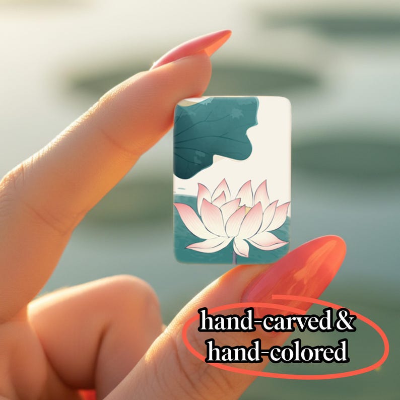 May include: A small rectangular tile featuring a hand-carved and hand-colored illustration of a pink lotus flower against a cream background with dark teal leaf accents. The surface displays the text hand-carved & hand-colored in black.