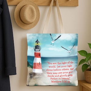 May include: A white tote bag featuring a lighthouse with red and white stripes, set against a blue sky and ocean. Two seagulls fly above. The bag includes the text "You are the light of the world...Matthew 5:14-16."