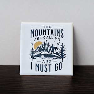 Peut inclure: Carreau carré blanc avec une scène de montagne noire et dorée et le texte "The Mountains Are Calling And I Must Go."