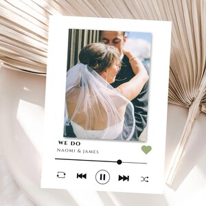 Puede incluir: Una tarjeta de boda blanca con una foto de una pareja abrazándose. La tarjeta presenta una interfaz de reproductor de música con el texto "WE DO" y los nombres "NAOMI & JAMES". También hay un icono de corazón verde.