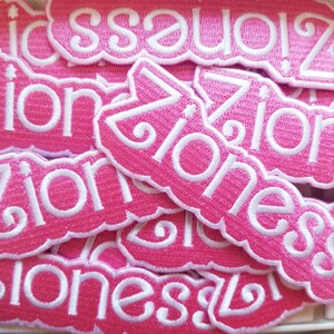 May include: A collection of pink embroidered patches with the word "Ziones" in white script. The patches have a scalloped edge and are stacked together, creating a textured surface.