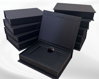 Puede incluir: Varias cajas rectangulares negras apiladas, algunas cerradas y otras abiertas. Una caja está abierta, revelando un interior empotrado con un recorte circular. Las cajas parecen ser de un material mate.
