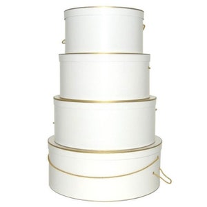 May include: A stack of four white round hat boxes with gold trim and handles. The boxes are nested, with the largest at the bottom and the smallest at the top. The handles are made of a gold-colored rope.