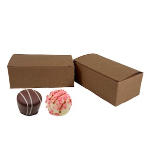 Peut inclure: Deux boîtes en carton brun avec couvercles, chacune contenant deux truffes au chocolat. Une truffe est au chocolat noir avec des rayures blanches, et l'autre est au chocolat blanc avec des pépites roses.