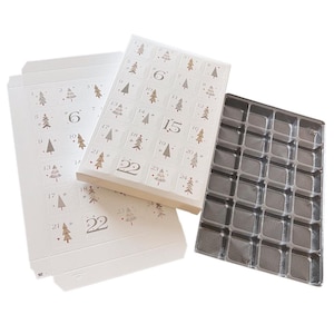 Puede incluir: Cajas blancas de calendario de adviento con números e ilustraciones de árboles de Navidad. Las cajas están abiertas, revelando una bandeja de plástico transparente con múltiples compartimentos. Los números 1-24 están impresos en las cajas.