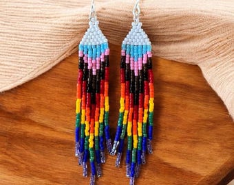 Puede incluir: Un par de pendientes largos colgantes con una sección superior de patrón en zigzag en azul claro, turquesa y rosa, que transiciona a hilos verticales de flecos de cuentas multicolores en negro, rojo, naranja, amarillo, verde y azul.