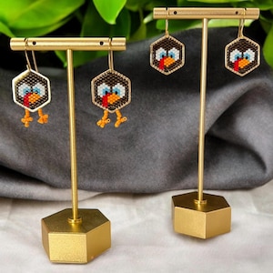 May include: Gold-toned earring display featuring two pairs of beaded turkey earrings. Each earring has a hexagonal shape with a brown background, a turkey face, and orange legs. The earrings are displayed on gold stands.