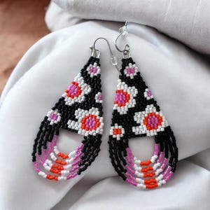 May include: A pair of black, white, pink, and orange beaded earrings with a teardrop shape and fringe. The earrings have a geometric pattern of circles and a dangling fringe.