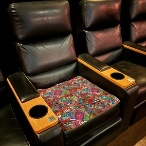 May include: Two black leather theater seats with wooden armrests and cup holders. The seat on the left has a colorful patterned cushion and the number "11" printed on the armrest. The seat on the right has a plain black cushion and the number "12" printed on the armrest.
