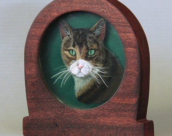 Puede incluir: Una pintura enmarcada de un gato atigrado con ojos verdes. El gato está mirando directamente al espectador. El marco es de madera oscura y tiene una parte superior redondeada.